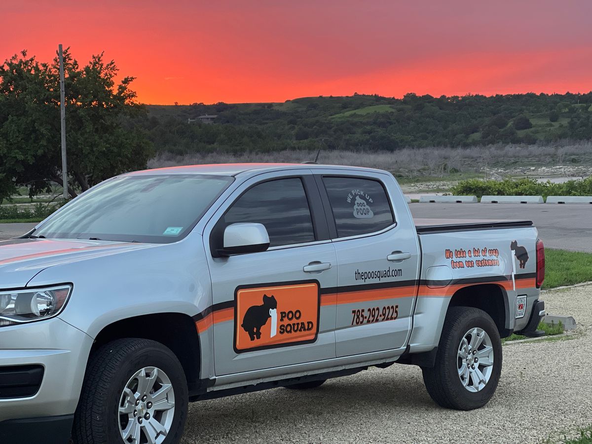 A branded Poo Squad pickup truck parked on a gravel drive at sunset, with an orange and pink sky behind low hills.