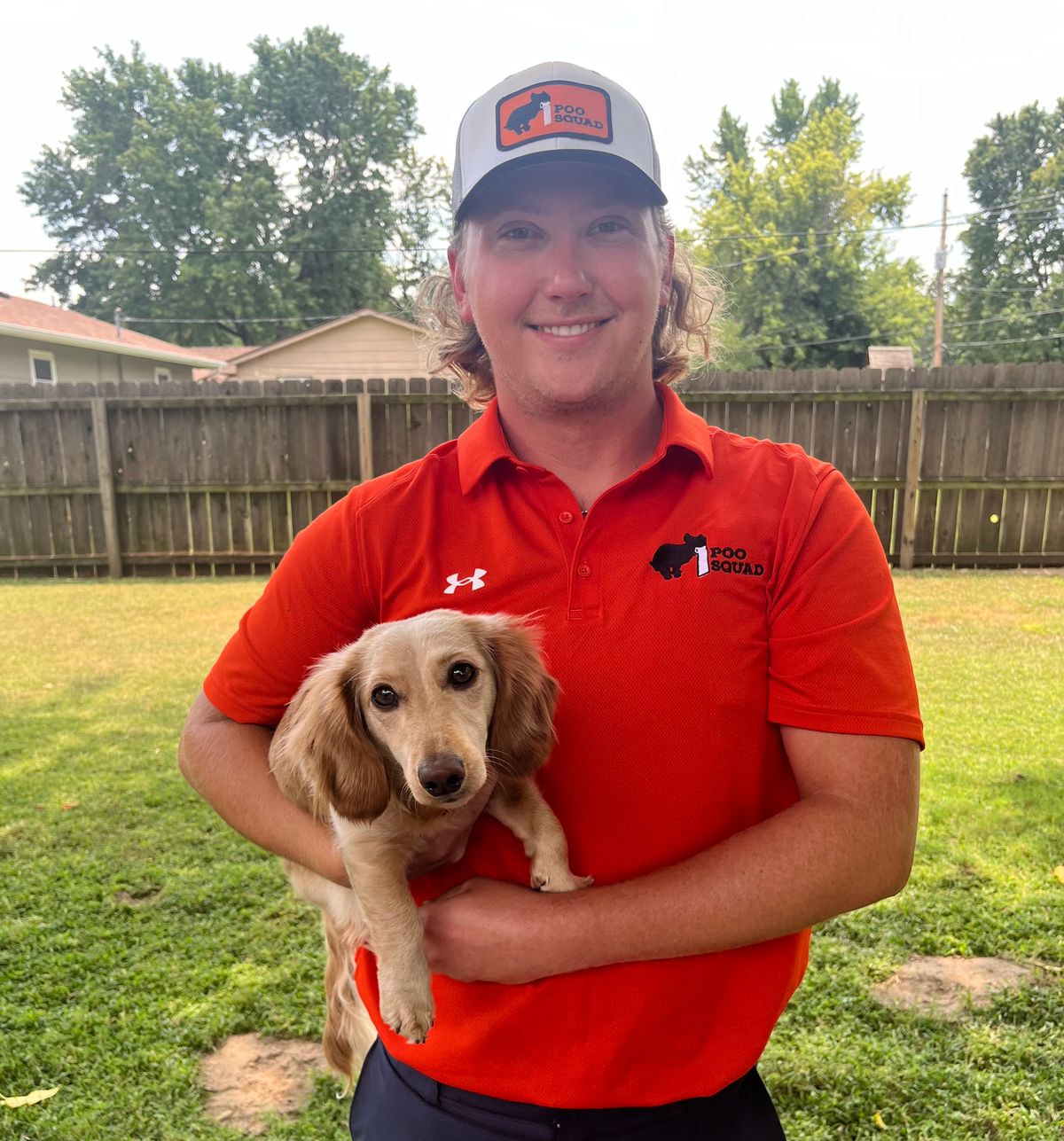 A Poo Squad crew member in an orange polo and branded cap standing in a fenced back yard, holding a small cream dachshund.
